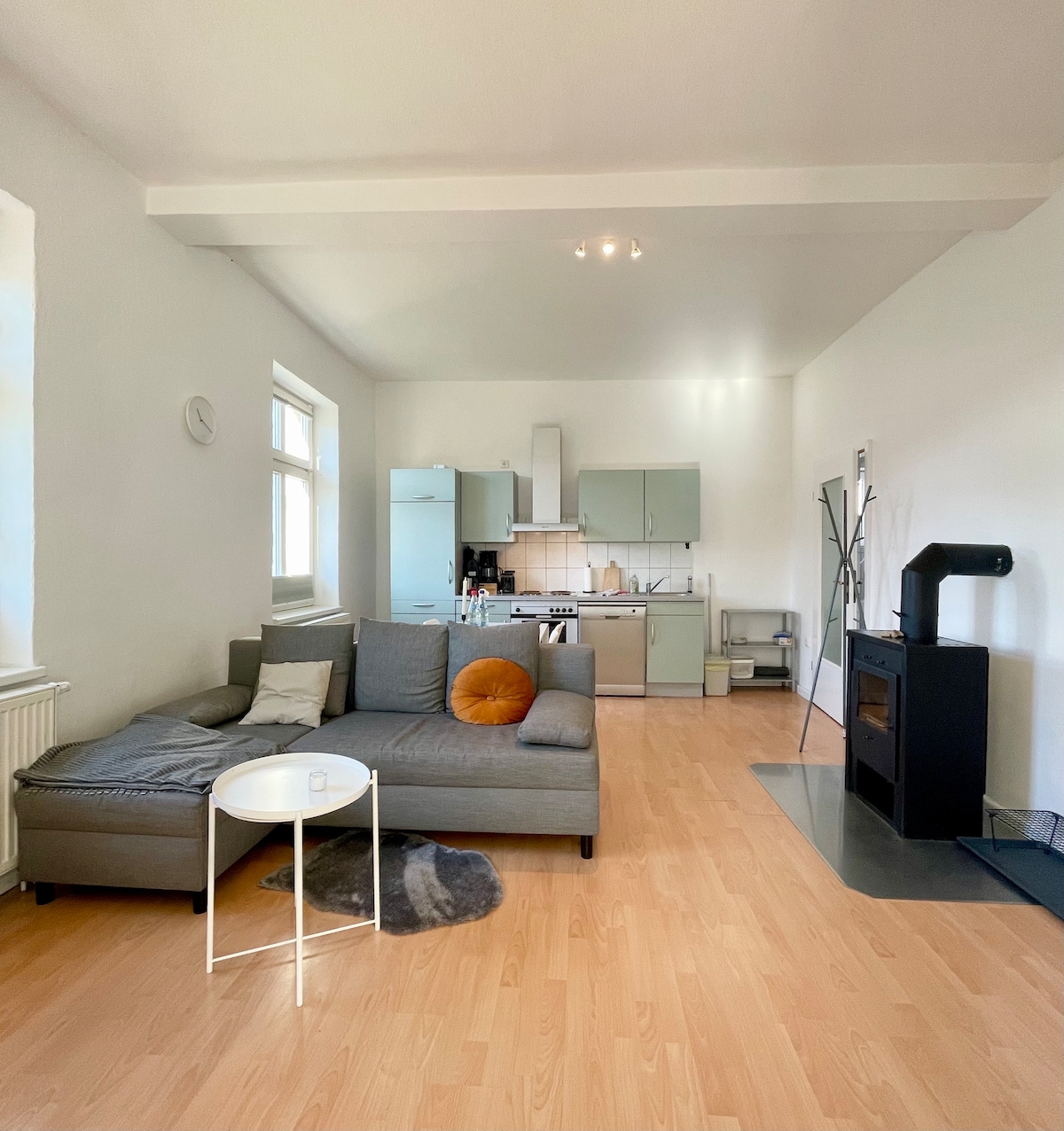 A spacious living area showcases a comfortable gray sectional sofa, complemented by an orange accent pillow. A small white table stands beside the sofa. The open kitchen features light-colored cabinets and is equipped with modern appliances. Natural light floods the room through large windows.