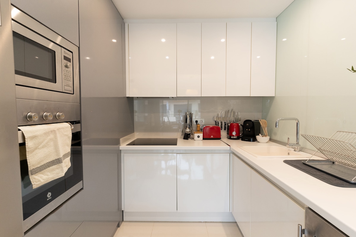 A modern kitchen is showcased, featuring sleek cabinetry in light gray and white. Stainless steel appliances, including an oven and a red kettle, are visible. A minimalist countertop is complemented by a dish rack and a sink, creating a clean and functional cooking space.