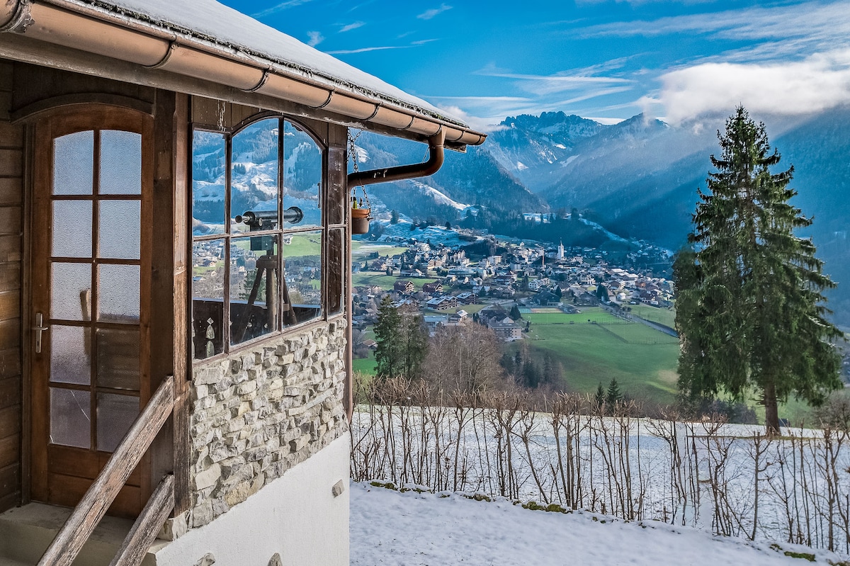 A wooden structure is showcased with a stone base, complemented by a charming door and large windows. The view features broad fields and a snow-covered landscape, with distant mountains and a village nestled in the valley, creating a serene and tranquil setting.