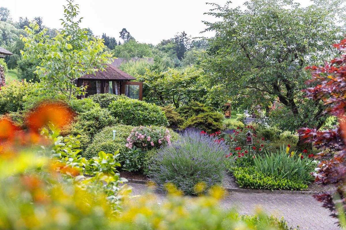 A lush garden is displayed, featuring dense greenery, flowering shrubs, and a variety of colorful plants. Paths wind through the vibrant landscape, leading towards cozy seating areas and a gazebo. The tranquil surroundings are framed by trees and the distant scenery of hills.