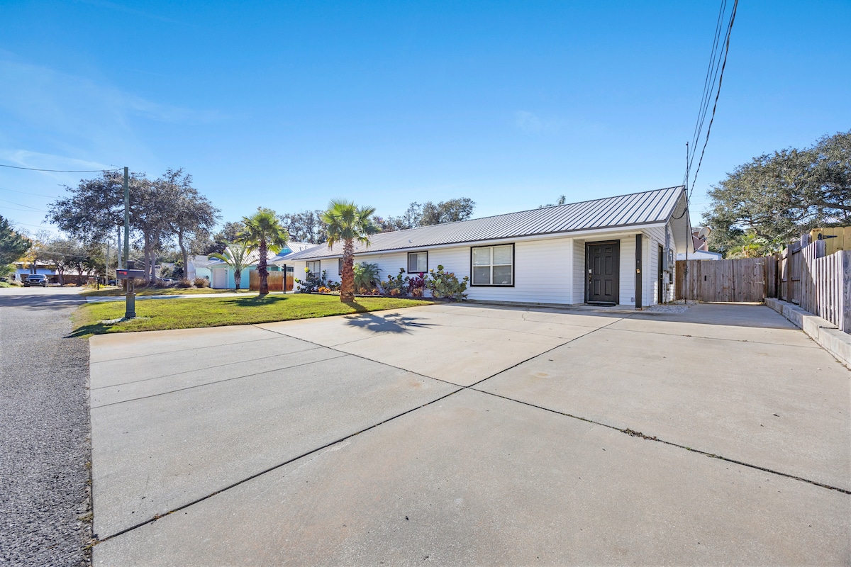 The exterior of the beach retreat is shown, featuring a welcoming single-story structure with a metal roof and a spacious concrete driveway. Lush greenery, including palm trees, enhances the outdoor space, which is surrounded by a peaceful neighborhood.