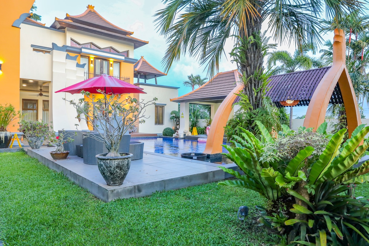 A landscaped garden features lush tropical plants surrounding a spacious patio area with outdoor seating. A large red umbrella provides shade over the seating. The scene includes a private pool and traditional pavilion, creating a serene outdoor setting.