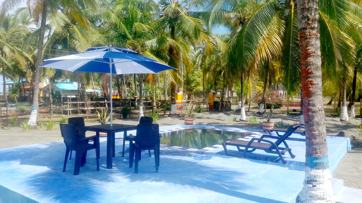 An outdoor pool area is surrounded by palm trees, featuring a shaded umbrella over a table with two chairs. A lounge chair is positioned near the edge of the pool, offering a quiet space for relaxation.