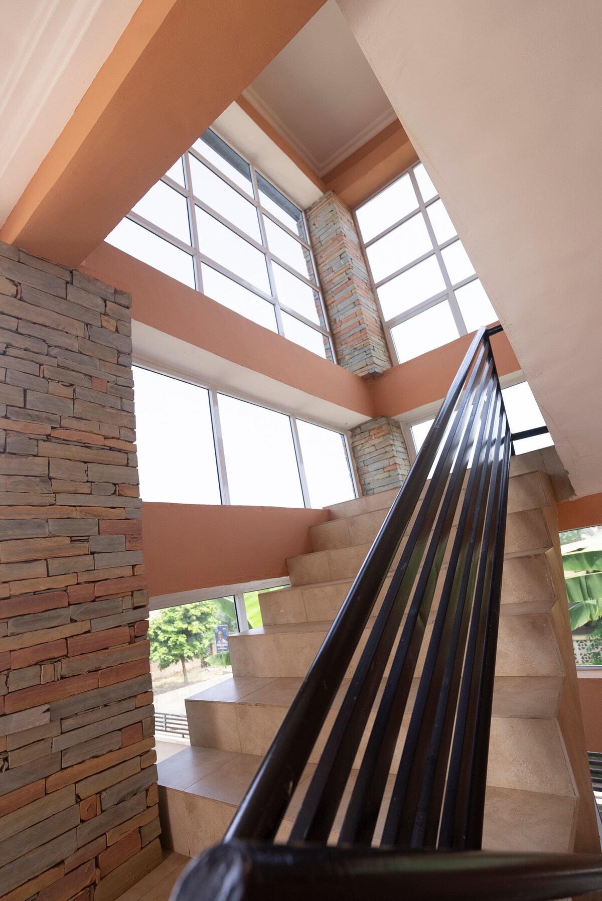 A staircase featuring a sleek, black railing ascends alongside large windows that allow natural light to fill the space. The walls combine stone and warm-toned plaster, creating an inviting and modern atmosphere. The greenery outside is visible through the expansive windows.