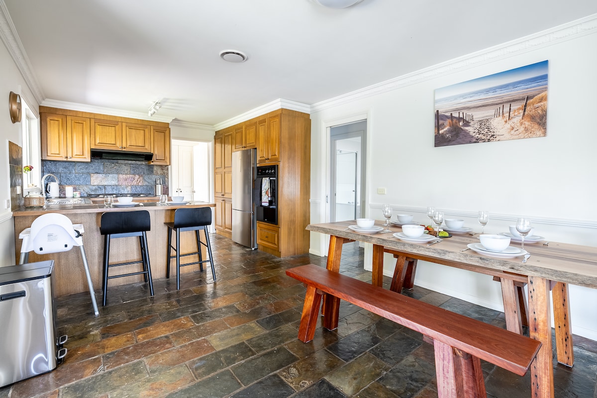 The kitchen and dining area feature stone flooring and wooden cabinetry, creating a warm ambiance. A large wooden dining table is set with glassware, while bar stools are positioned at the kitchen counter. Appliances, including a refrigerator and a wall oven, are visible, with natural light flowing through nearby doors.