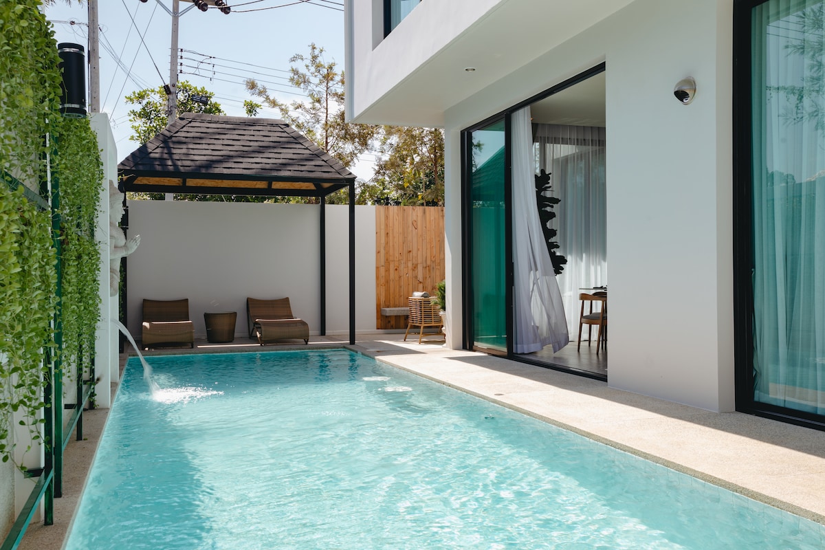 A tranquil private pool reflects the sunlight and is surrounded by a stylish patio area. Two lounge chairs provide a comfortable space for relaxation, while a small table adds functionality. Large glass doors invite views of the interior living space, enhancing the open design.