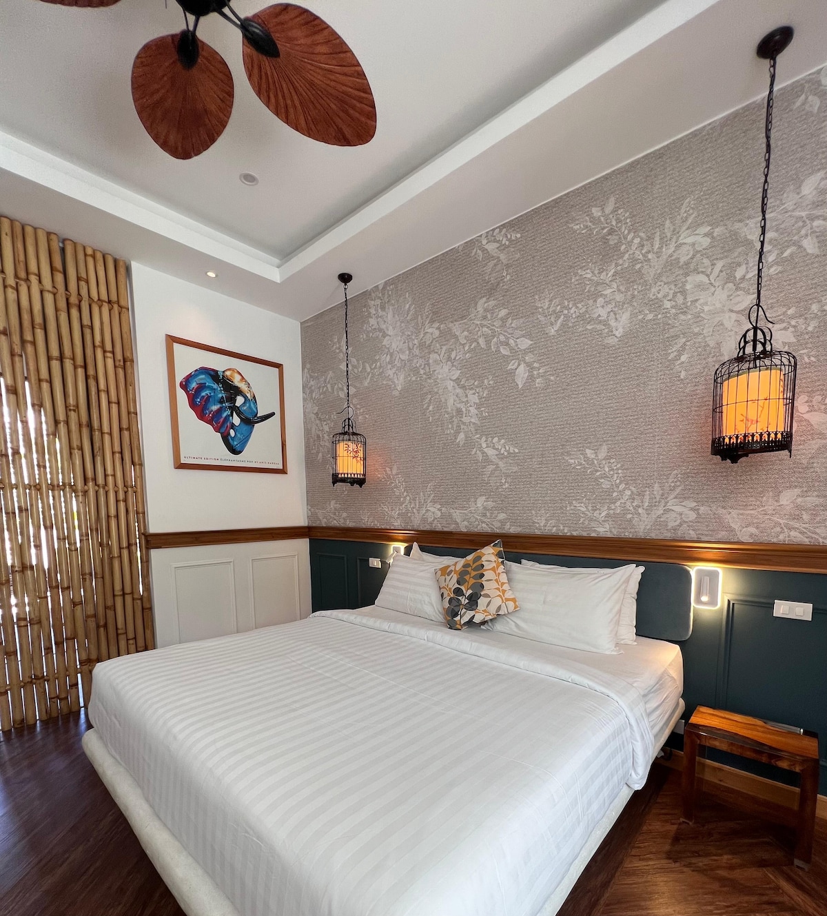 A cozy bedroom features a spacious bed with crisp white linens. Artistic wall decor includes a framed piece and patterned wallpaper. Warm light is provided by hanging lanterns, while a bamboo screen adds a touch of natural texture. Wooden accents are present in the furniture.