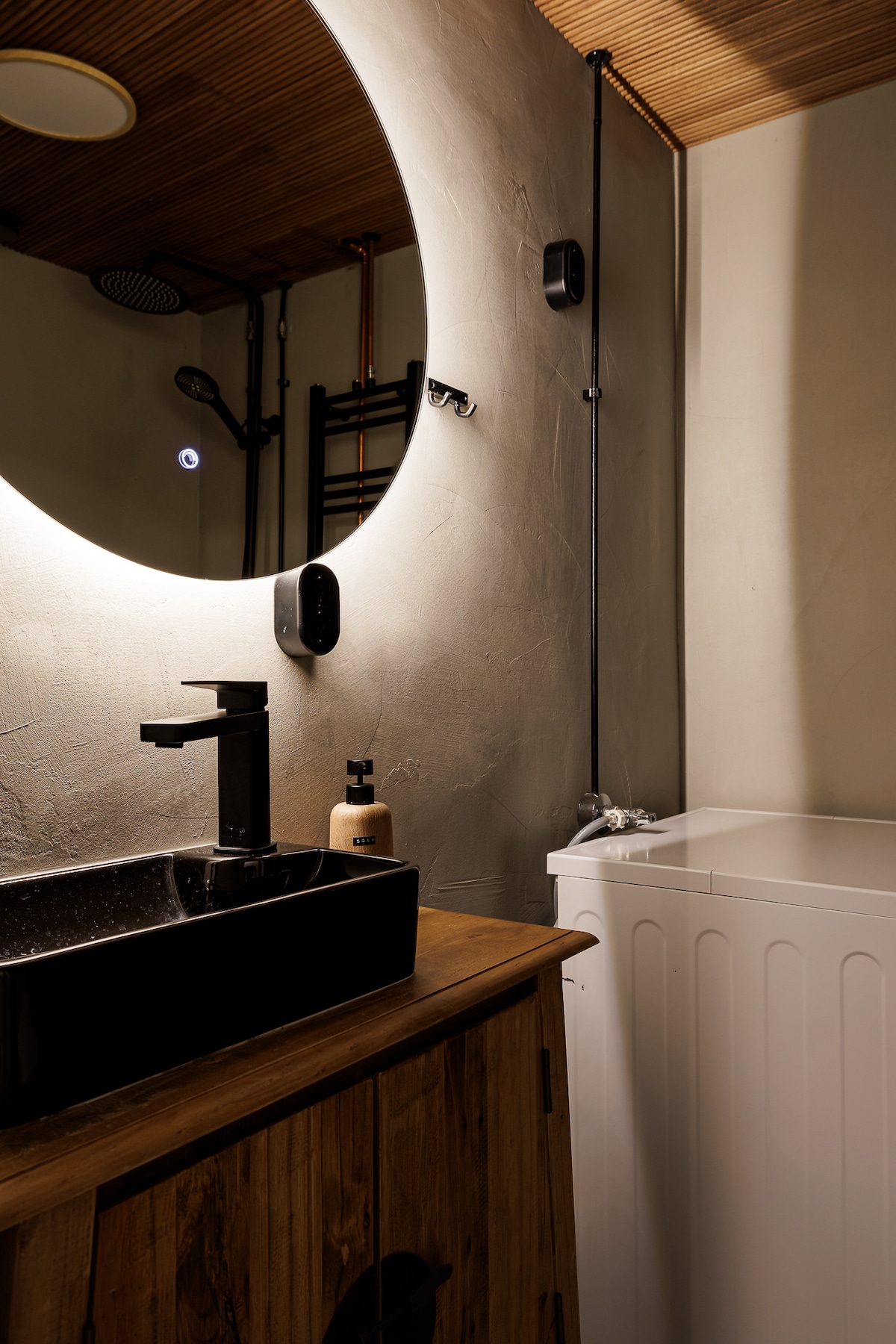 The bathroom features a circular mirror illuminated by soft light, providing a modern aesthetic. A sleek black sink is positioned atop a wooden vanity, complemented by a neutral-walled backdrop. A compact washing machine is visible, integrating functionality into the stylish design.