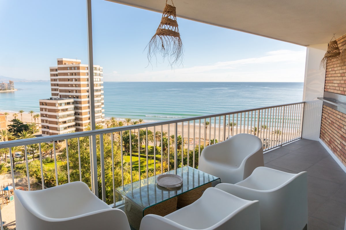 A spacious terrace features a glass table surrounded by white armchairs. The view includes a sandy beach and calm sea, complemented by palm trees. Buildings stand in the background, and sky meets the water on the horizon, creating a serene coastal scene.