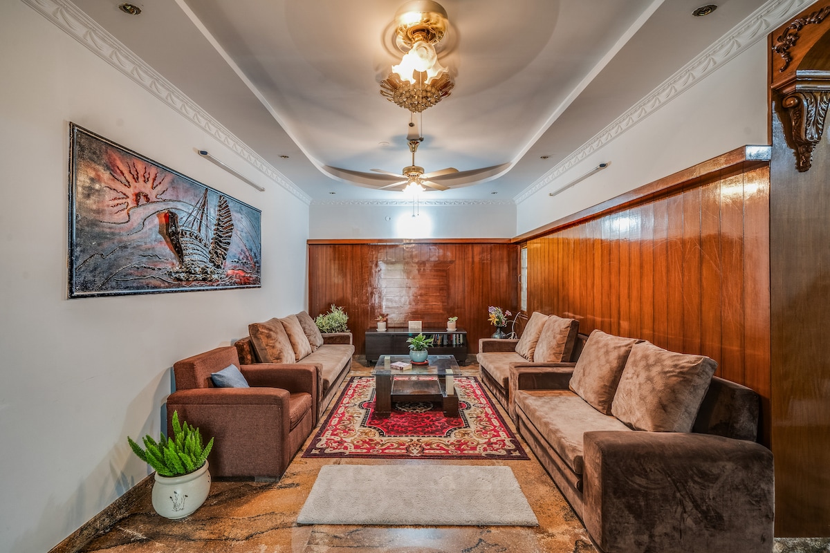 A spacious hall features soft lighting and elegant furnishings, including multiple plush sofas arranged around a central coffee table. A richly patterned area rug enhances the space, complemented by wooden panels and decorative plants. A wall-mounted artwork adds a touch of color to the inviting atmosphere.
