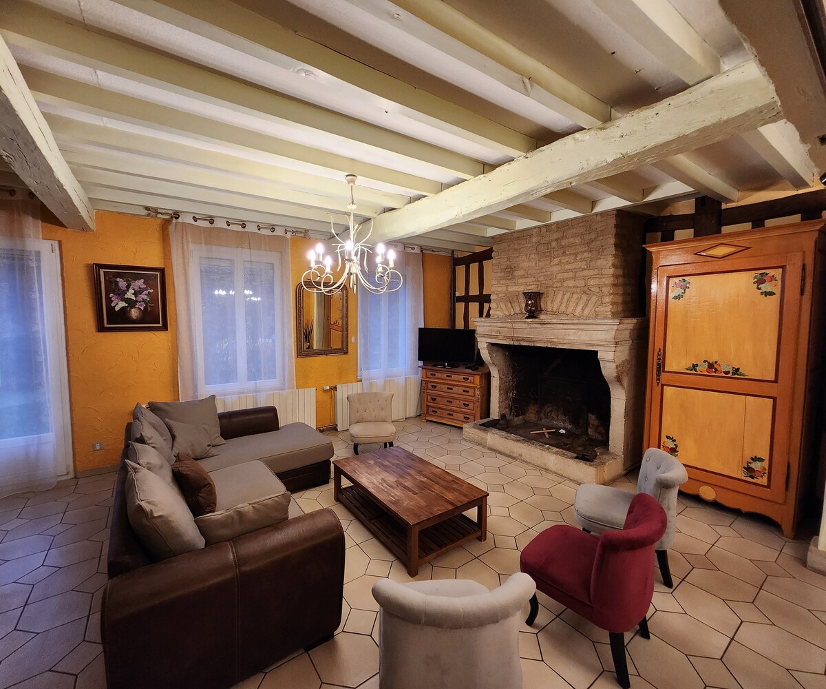 The living area features a stone fireplace and a large seating arrangement that includes a mix of sofas and armchairs. A wooden coffee table is centrally placed, and a vintage cabinet adds character. Two windows provide natural light, complemented by soft drapery.