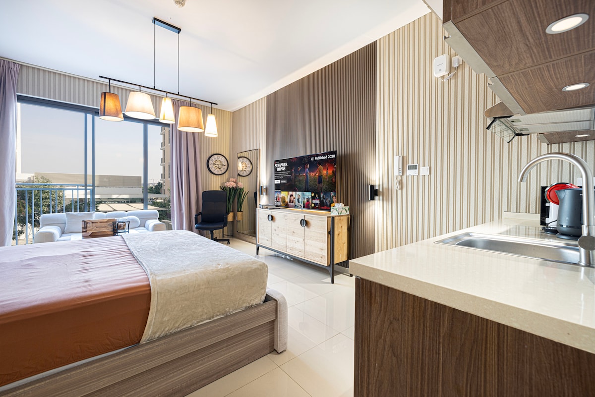 A stylish studio space features a spacious bed adorned with neutral bedding. Large windows allow natural light to fill the room, showcasing views of the outdoor area. Contemporary furniture, including a seating area and a media console, complements the modern design.