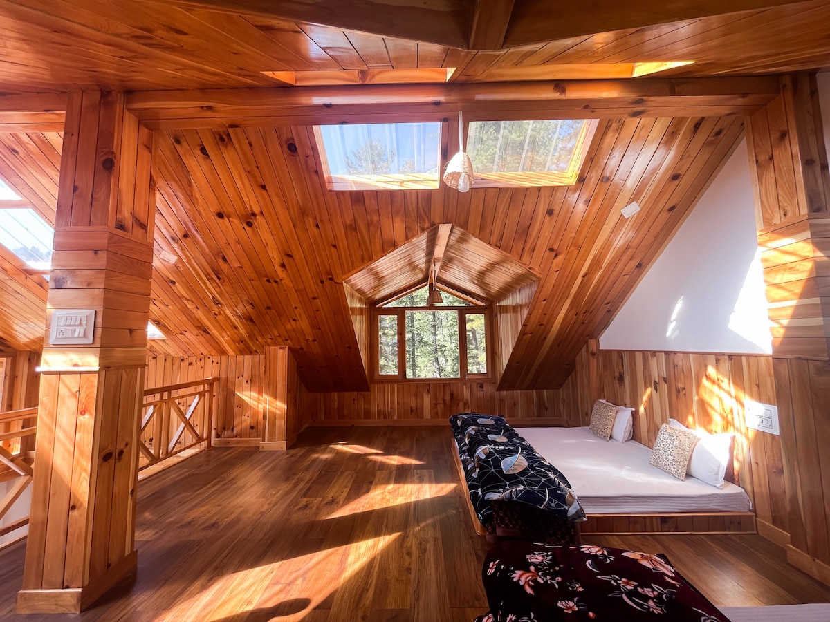 The attic space features wooden walls and ceiling beams, creating a cozy atmosphere. Skylights allow natural light to illuminate the area. A spacious mattress is positioned on one side, adorned with decorative cushions, while large windows provide views of the surrounding nature.