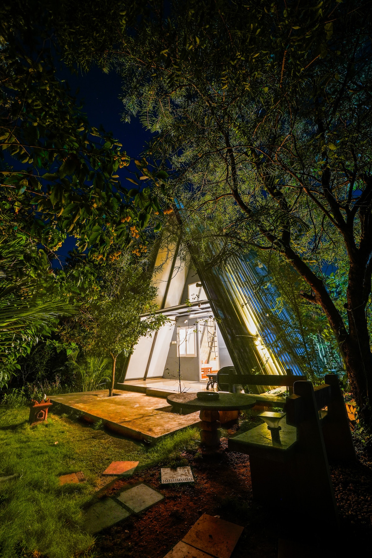 The cabin house features a unique A-frame design, illuminated at night. Surrounding greenery frames the structure, with stone pathways leading to a small seating area. Soft lighting highlights the cabin's architectural angles against a dark sky.