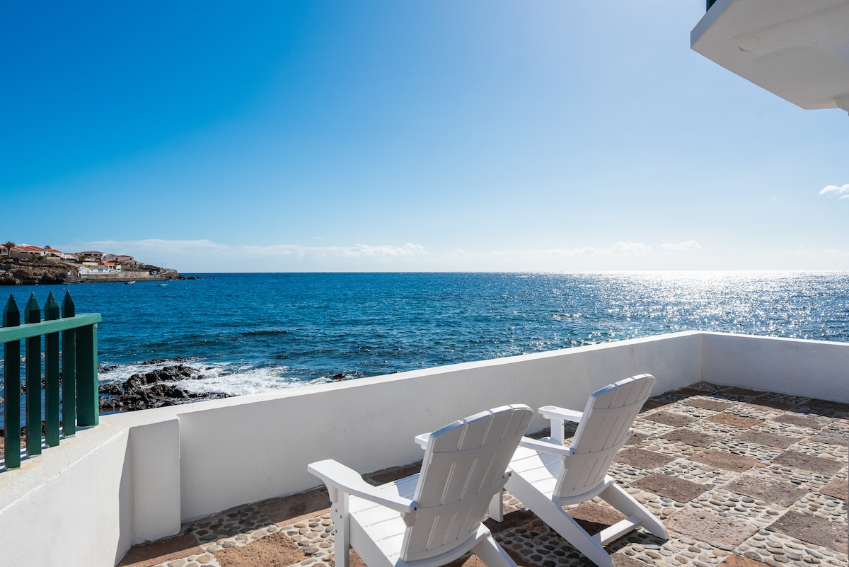 A private stone terrace overlooks the serene ocean, featuring two white lounge chairs positioned for relaxation. The sparkling water reflects sunlight under a clear blue sky, creating a tranquil outdoor space ideal for enjoying the surrounding coastal views.