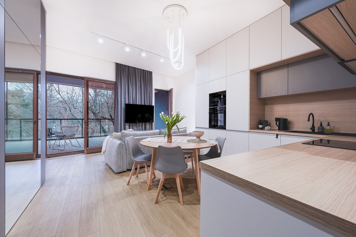 The modern kitchen and dining area features a round table surrounded by four grey chairs. Light wood flooring complements the cabinetry. A large glass door leads to the terrace, providing a view of the greenery outside. A stylish light fixture hangs above the table.