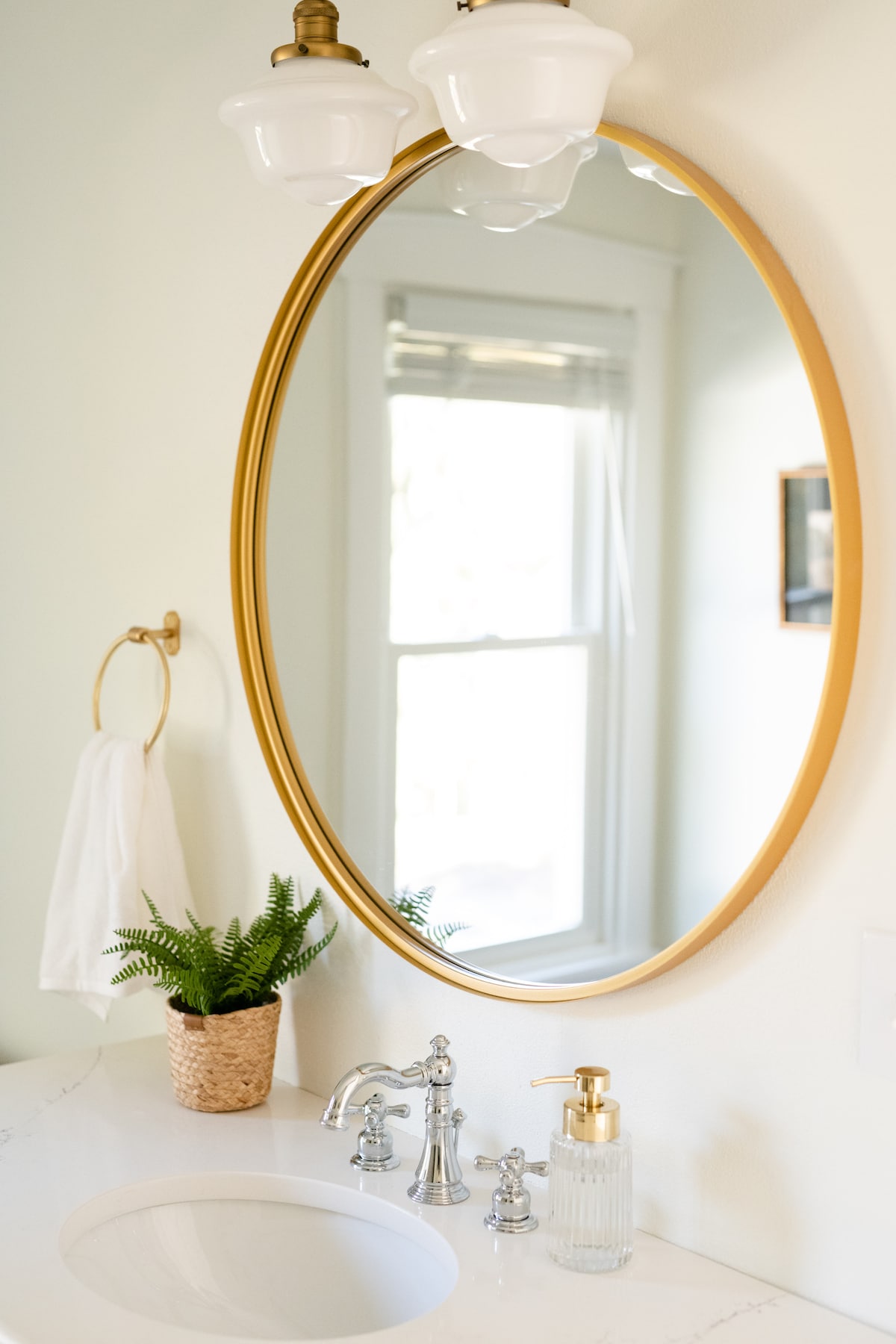 A round mirror with a gold frame reflects a well-lit bathroom setting. Below, a white sink is accompanied by a modern faucet and a soap dispenser. A potted fern adds a touch of greenery to the countertop, while a towel hangs on a gold ring nearby.