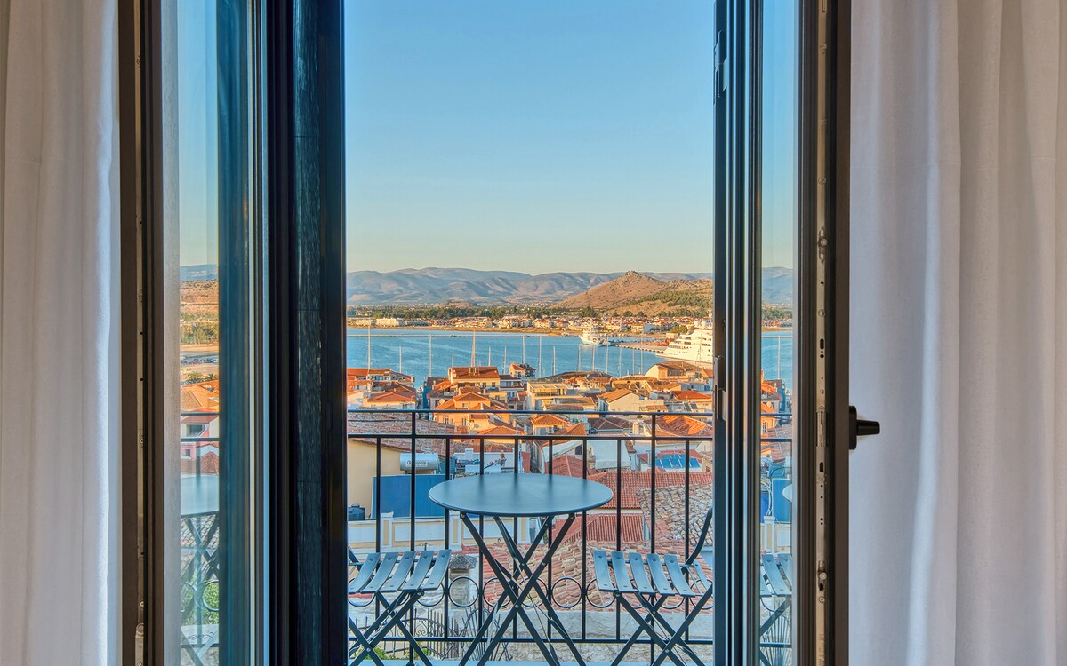 A wide view is presented from an open window, showcasing a landscape of Nafplio's rooftops below. A small round table and chairs are situated on the balcony, overlooking the serene waters of the Argolic Gulf and distant hills under a clear sky.
