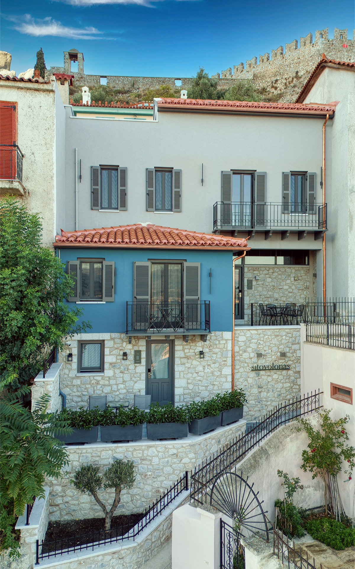 The exterior showcases a two-story building featuring a blend of stone and modern architecture. Large windows and balconies open to a neatly landscaped courtyard. Above, the historic fortress wall can be seen against a clear sky, enhancing the connection to Nafplio's heritage.