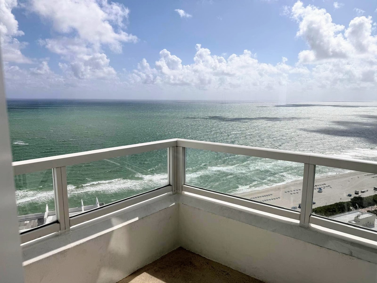 A furnished balcony offers a panoramic view of the ocean and shoreline. Soft waves are visible beneath a bright sky with scattered clouds, enhancing the serene coastal ambiance. The balcony's glass railings provide unobstructed views of the coastline and water's edge.