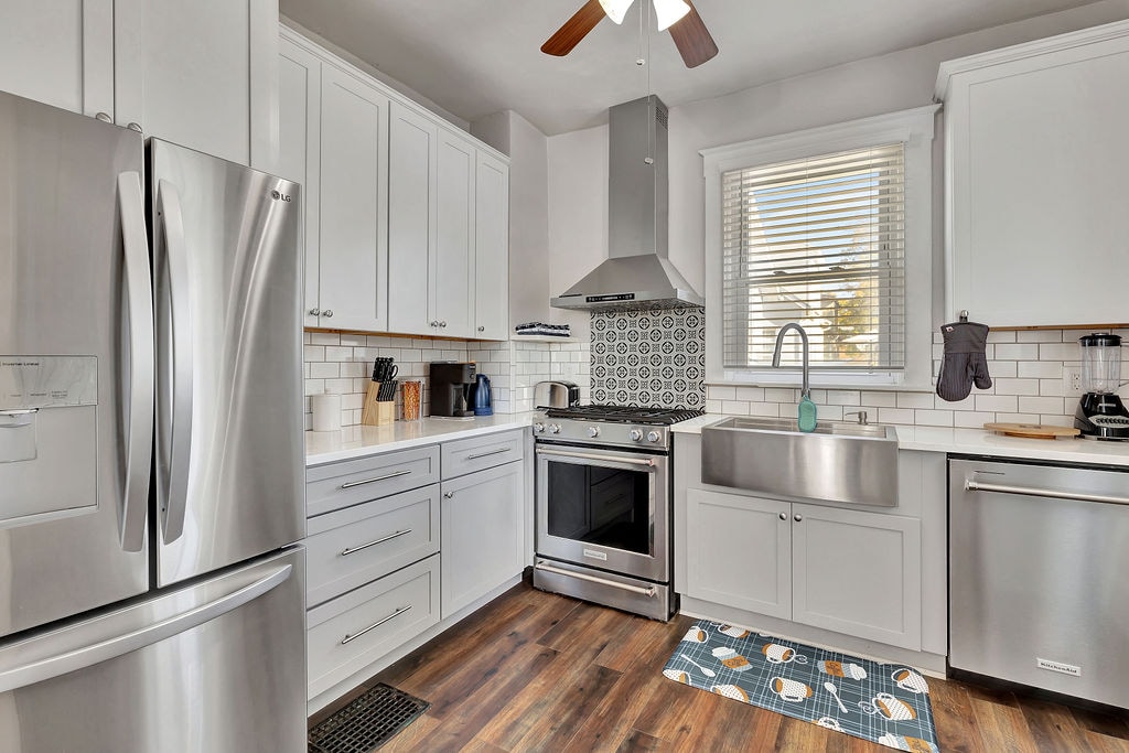 The modern kitchen features stainless steel appliances, including a refrigerator and a range. White cabinetry is complemented by a detailed backsplash. A large sink is positioned under a window, allowing for natural light. A textured rug adds color to the wooden floor.