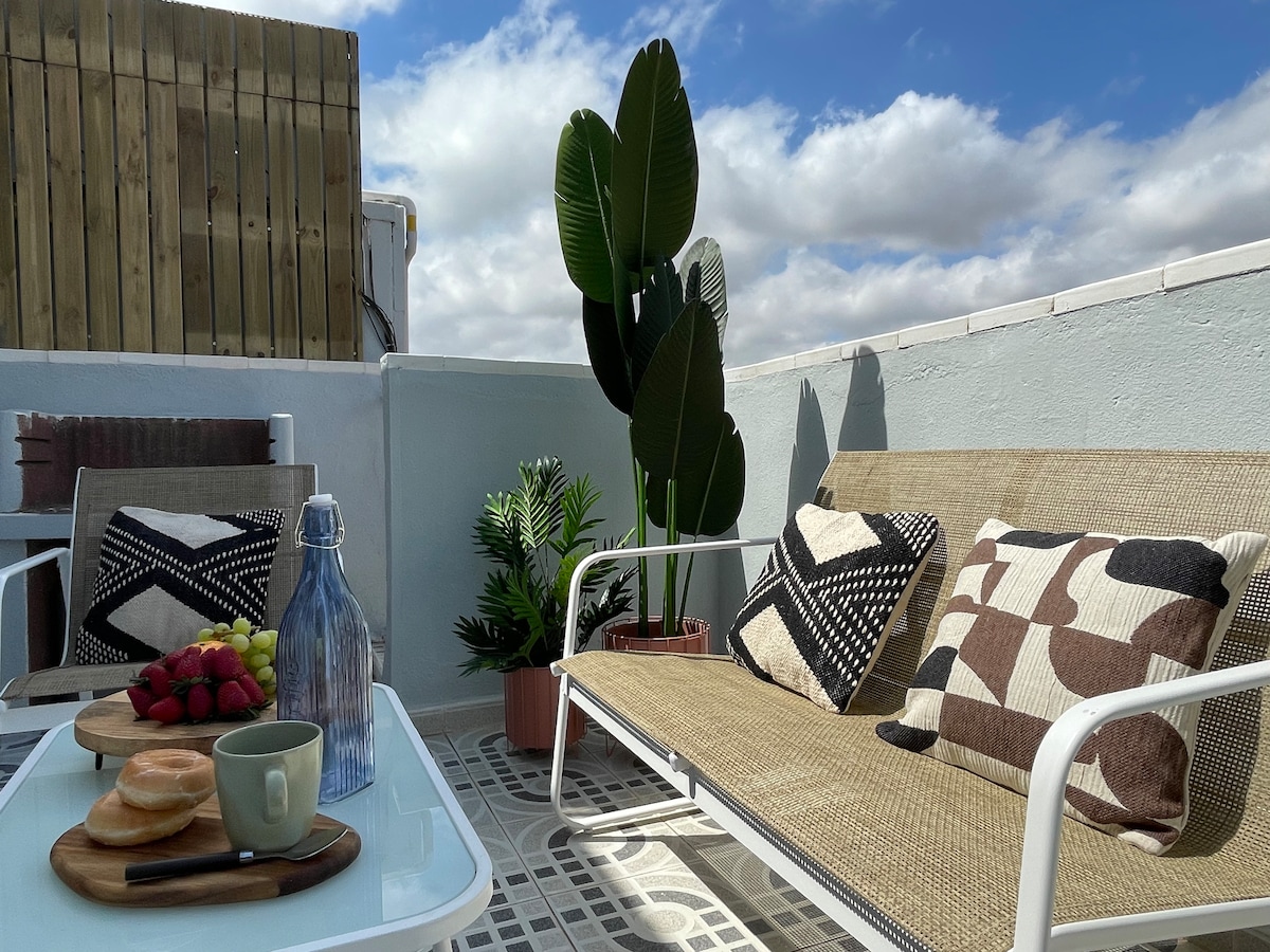 A sunny terrace is showcased, featuring two lounge chairs adorned with patterned cushions. A small table displays a bottle of water, a cup, fresh fruit, and pastries. Potted plants add greenery against a backdrop of a clear sky with scattered clouds.