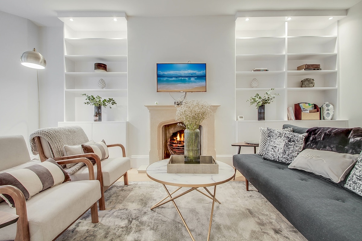 An inviting living area features a modern fireplace framed by white built-in shelves, displaying decorative items. A round coffee table sits at the center, complemented by a mix of comfortable seating, including a grey sofa and wooden armchairs. Soft lighting enhances the cozy ambiance.
