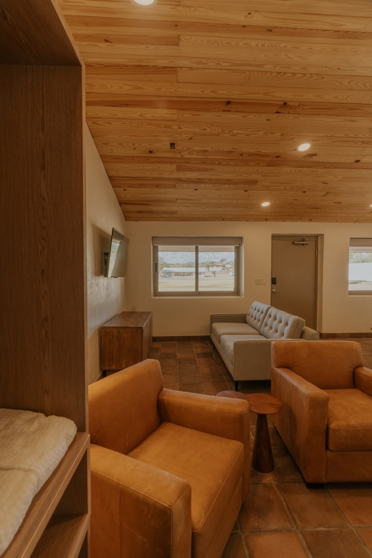 A spacious living area is characterized by a warm wood ceiling and earthy-toned tile flooring. Two orange chairs are positioned near a small side table. A light-colored couch offers additional seating, while natural light enters through a window adorned with simple window treatments.