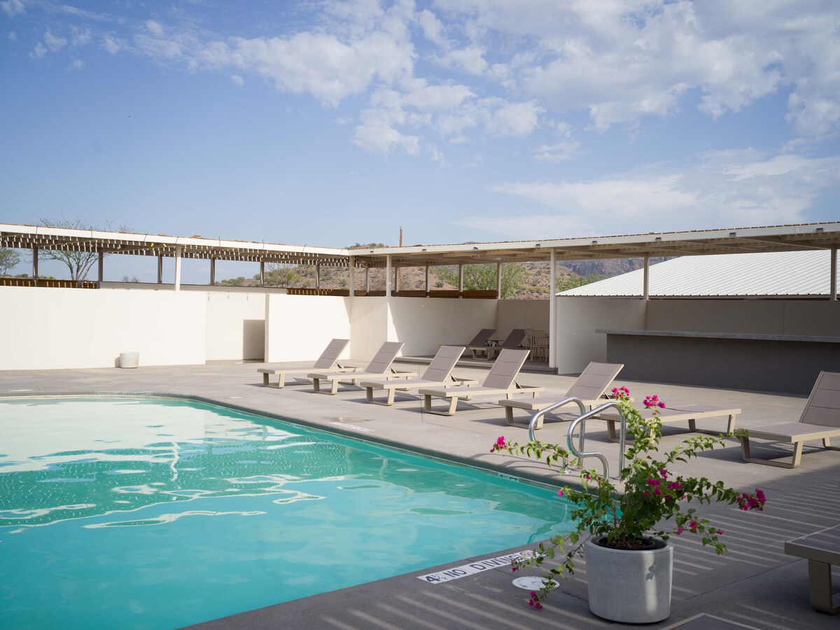 A serene pool area is highlighted by comfortable lounge chairs arranged along the deck. A flowering plant in a pot adds a touch of nature by the poolside. A shaded seating area is visible in the background under a trellis structure.