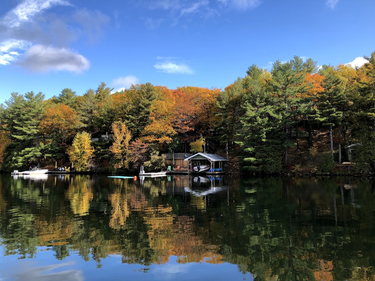 A serene lake view is presented, with autumn foliage reflected on the calm water. A cottage and boathouse are nestled among vibrant trees, while a dock extends into the lake. Soft clouds drift across a bright blue sky, enhancing the tranquil atmosphere.