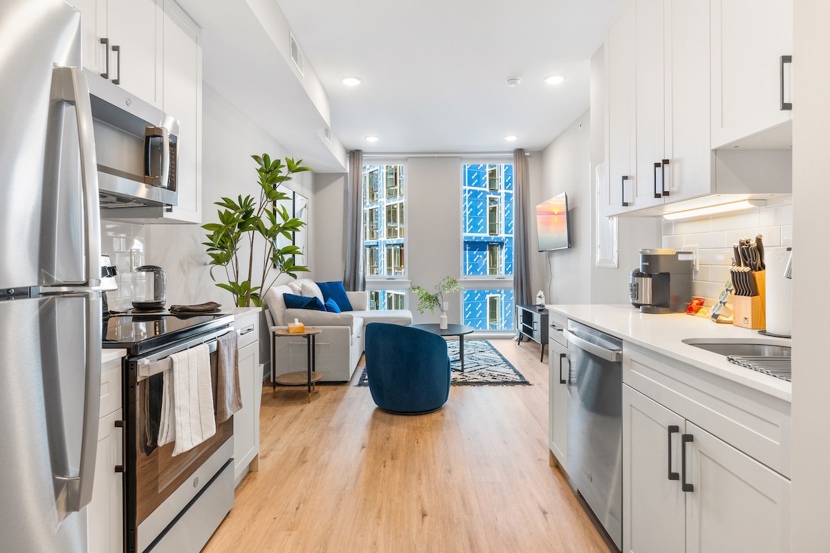 The image showcases a modern kitchen with stainless steel appliances and granite countertops. An open-concept living area features a sectional sofa, a coffee table, and a 50-inch Smart TV. Sunlight enters through large windows, accentuating the inviting and spacious atmosphere.