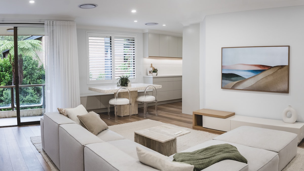 A modern living area is presented, featuring a spacious L-shaped sofa adorned with neutral-colored cushions. A minimalist wooden table sits on a textured rug, while a well-equipped kitchen is visible in the background. Natural light filters through the sliding glass doors, enhancing a calming atmosphere.