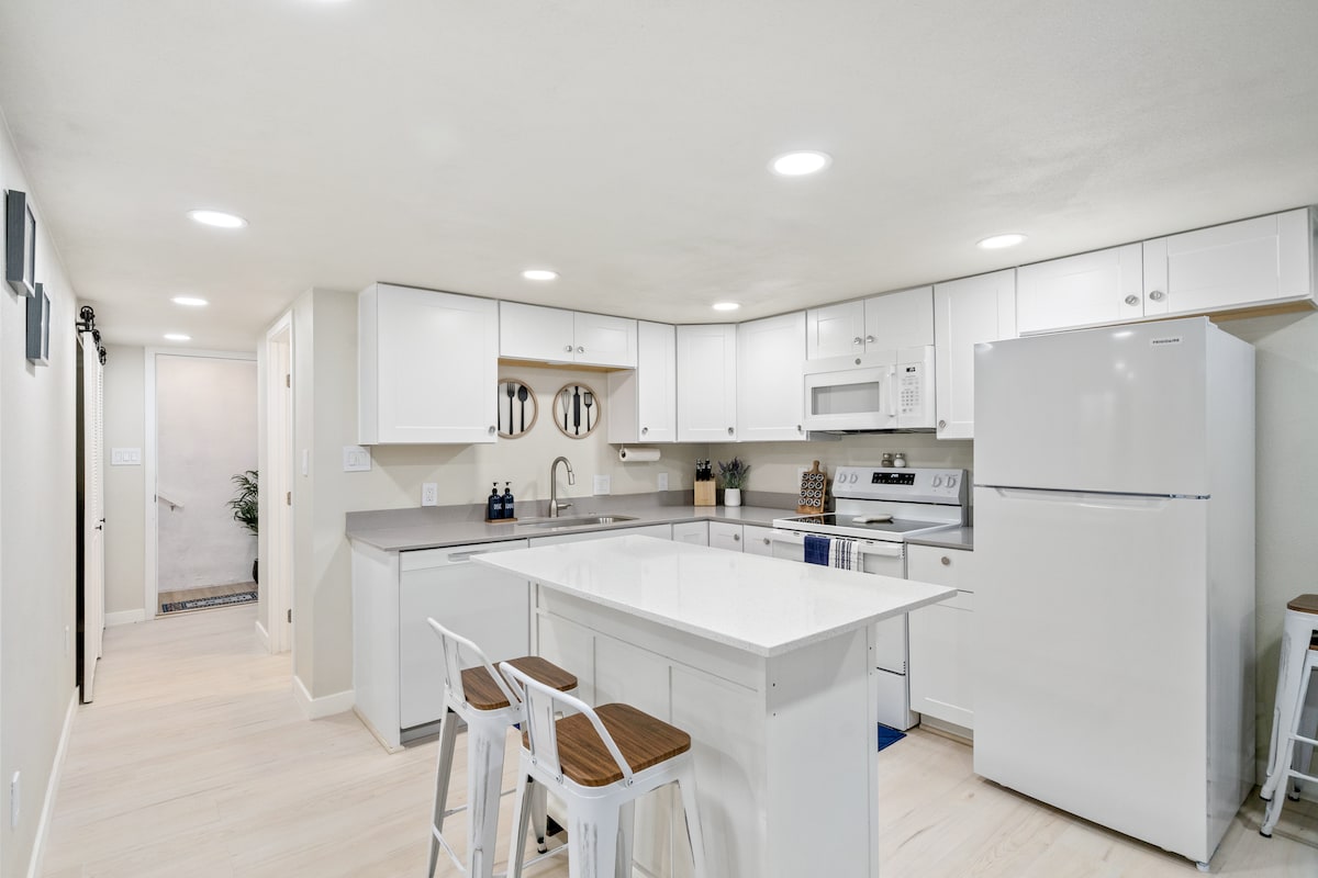 A modern kitchen features sleek white cabinetry and a central island with two wooden stools. Stainless steel appliances include a refrigerator and microwave, while ample countertop space enhances functionality. Bright overhead lighting illuminates the area, creating an inviting environment for cooking and dining.