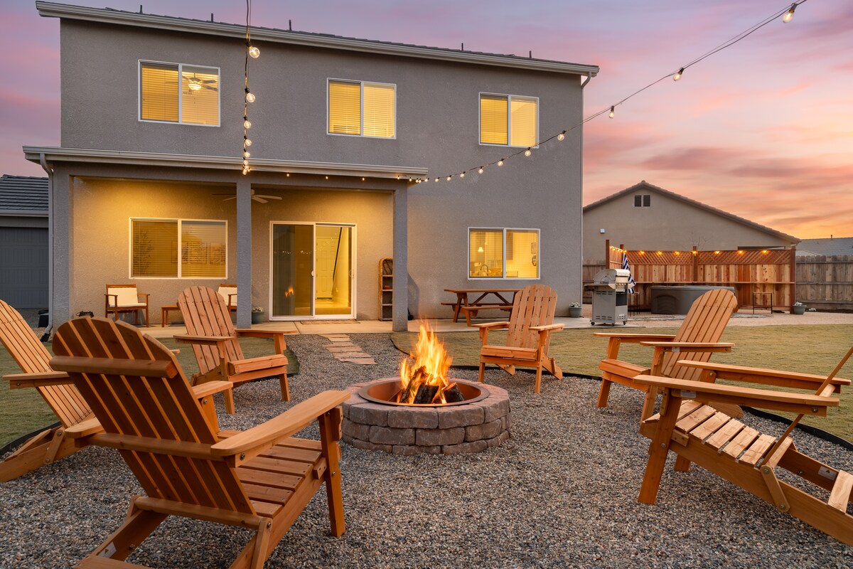 An outdoor space features a circular fire pit surrounded by wooden Adirondack chairs, set on a gravel area. The two-story home is visible in the background, with multiple windows reflecting the warm light of sunset. String lights are hung above, enhancing the ambiance.