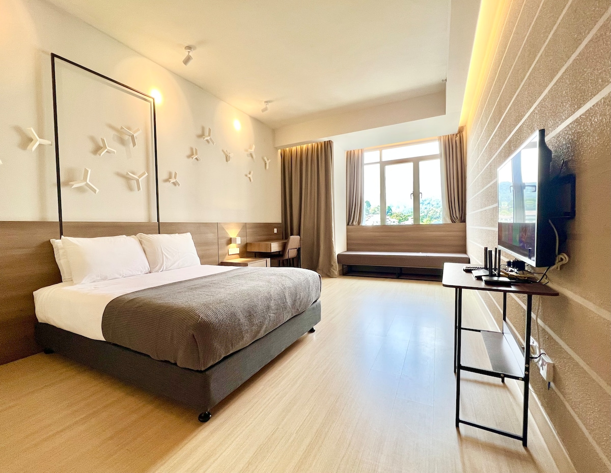 A spacious room features a comfortable queen bed with a neutral-toned comforter. Wooden accents provide warmth, while wall decorations consist of abstract shapes. A large window offers natural light and views, alongside a seating area and a 40" LCD TV mounted on the wall.