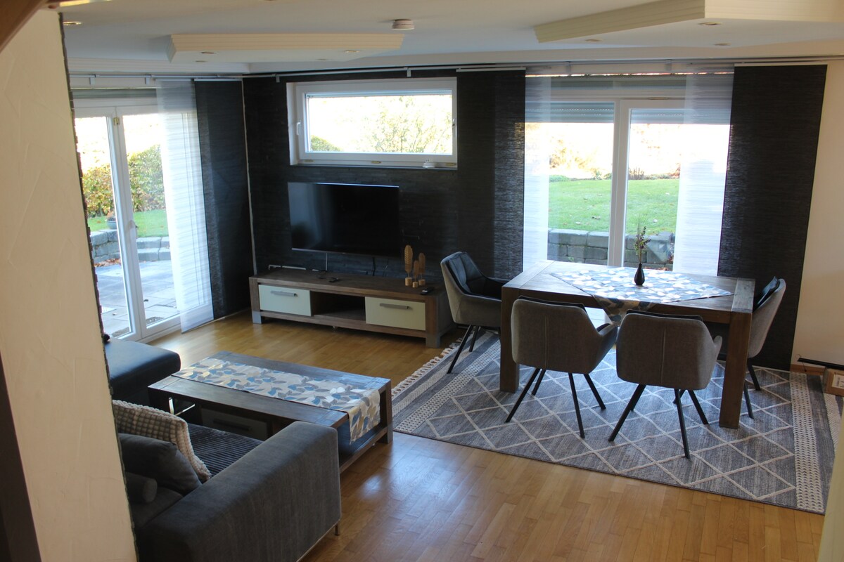 An open living area features a comfortable sofa and a dining table with four chairs. Large windows allow natural light, showcasing views of the outside landscape. A television is mounted on a media console, and a geometric rug adds texture to the wooden flooring.
