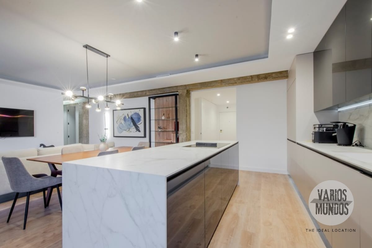 A modern gourmet kitchen features sleek marble countertops and a central island, complemented by dark cabinetry. A dining area is visible with stylish seating for six. Soft lighting illuminates the space, enhancing the clean lines and contemporary design, while a wall-mounted TV adds a touch of convenience.