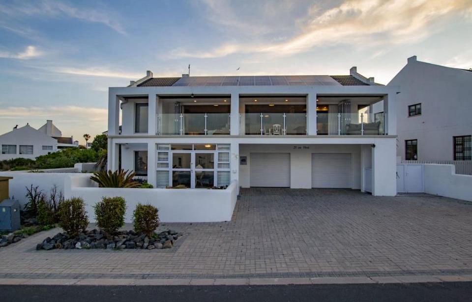 The exterior of the villa showcases a modern two-story structure with large glass balconies and a clean white facade. A paved driveway leads to a double garage, while landscaped gardens frame the entrance. The peaceful atmosphere is complemented by the surrounding residential area.