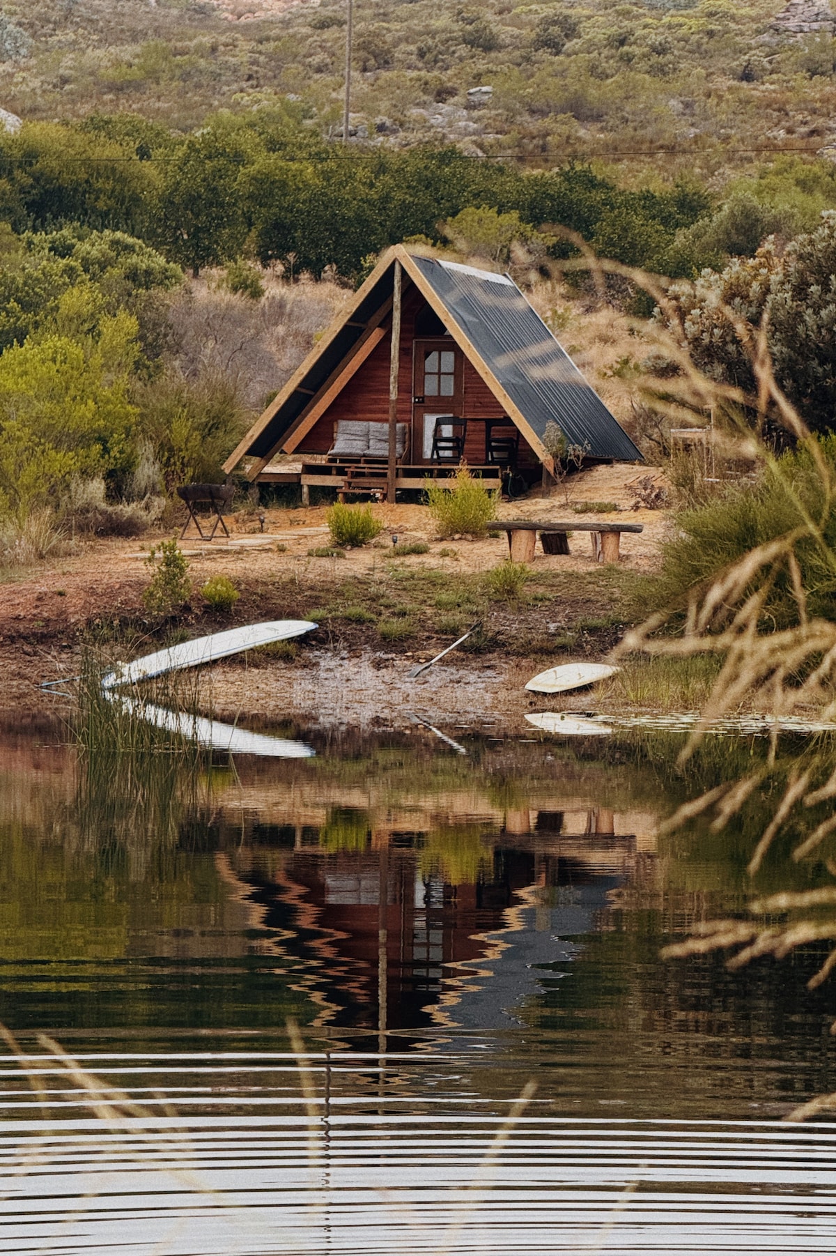 A serene A-frame cabin is nestled beside a calm dam, with wooden decking and simple outdoor furniture visible. The reflection of the cabin in the water creates a harmonious scene, framed by lush greenery and gentle ripples on the surface.