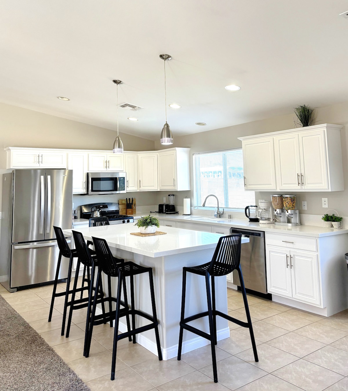 A modern kitchen is equipped with stainless steel appliances, including a refrigerator and microwave. A central island with four black bar stools offers seating. Bright natural light streams through the window, enhancing the clean lines of the white cabinetry and countertops.