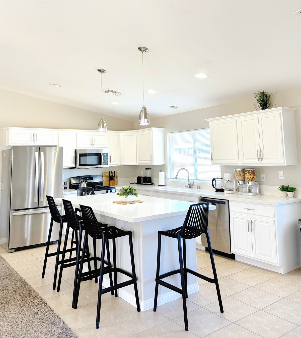 A modern kitchen features white cabinetry and stainless steel appliances. A spacious island with seating for four black chairs invites casual gatherings. Ample natural light illuminates the room, highlighting the clean design and well-organized space ready for meal preparation.