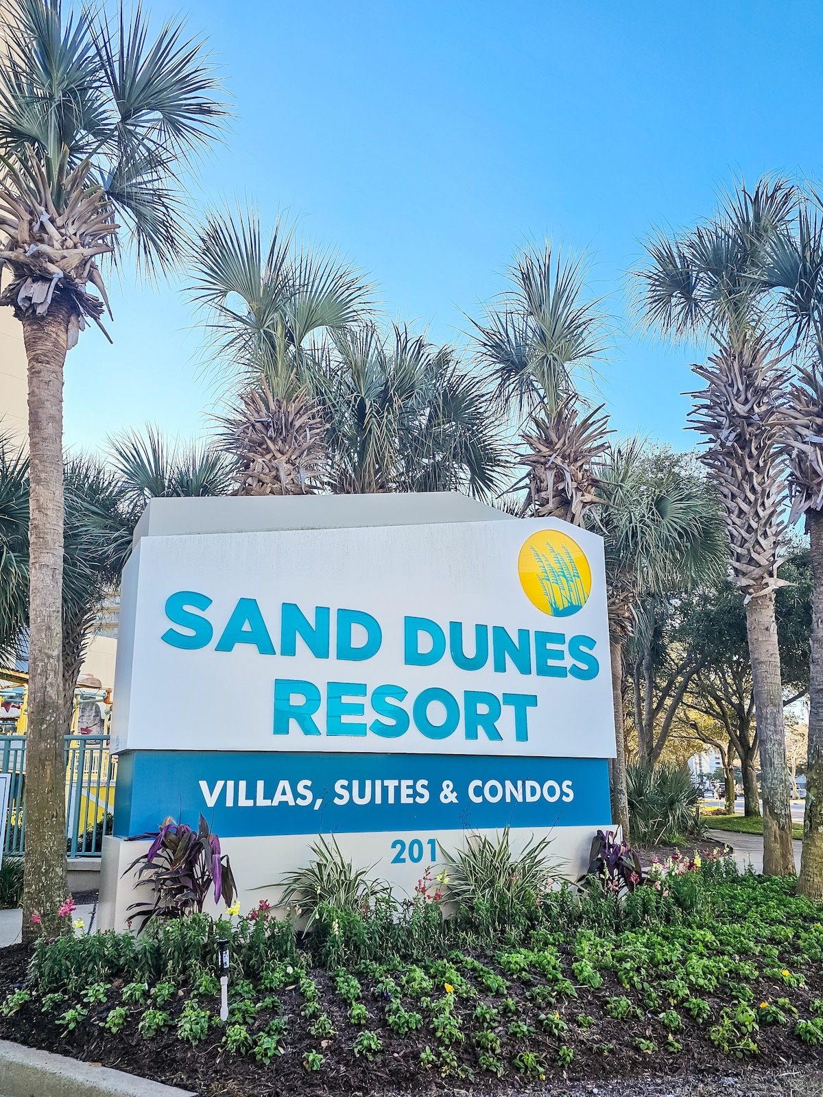 The entrance sign of Sand Dunes Resort is prominently displayed, featuring the name in bold blue letters. Surrounding palm trees and greenery enhance the welcoming feel. A clear blue sky is visible above, contributing to the pleasant outdoor atmosphere.