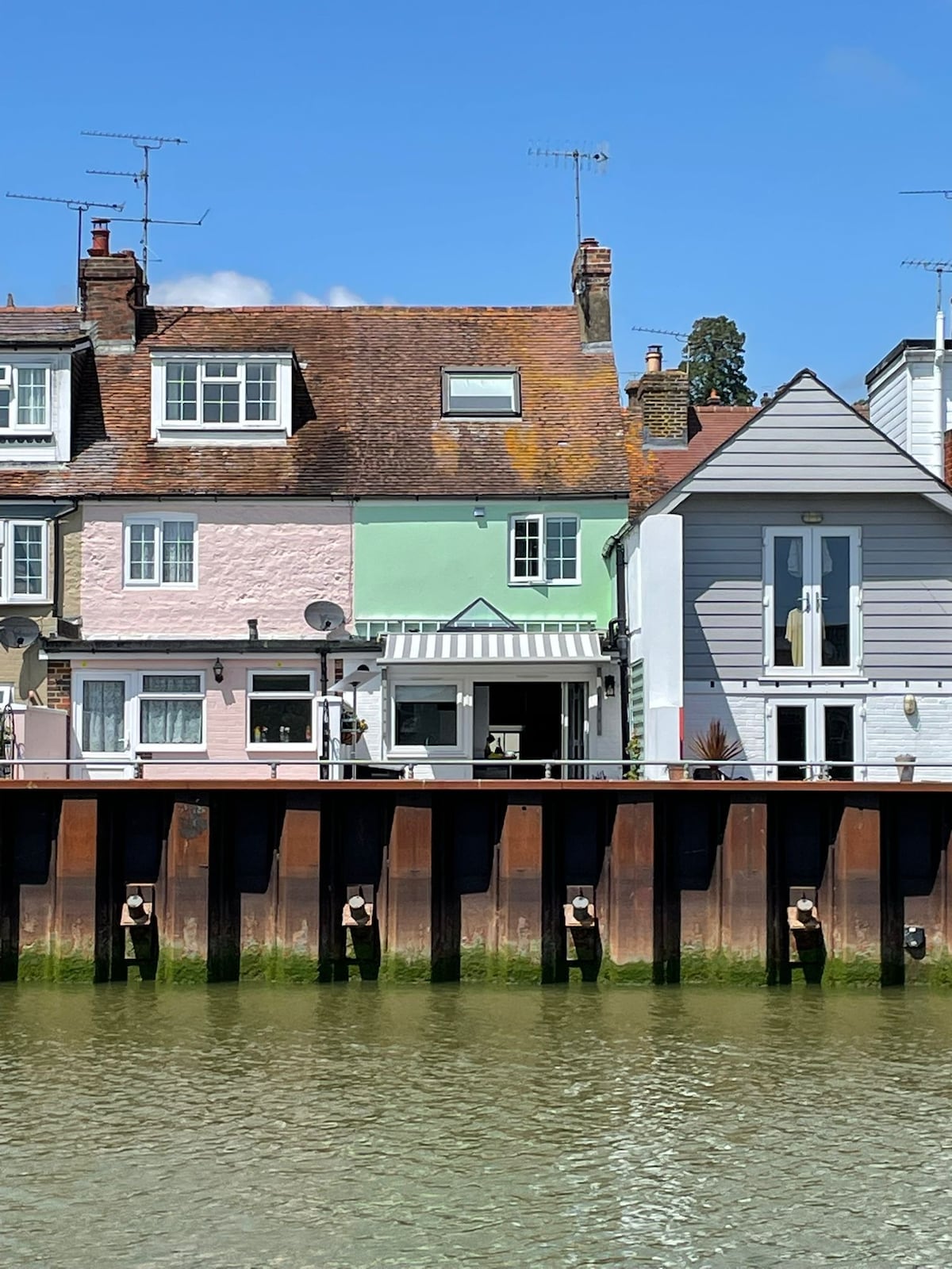 A charming row of 18th century cottages are seen along the waterfront. The buildings feature a mix of pastel colors, including soft pink and green. A modern terrace with large windows offers views of the river, enhancing the serene riverside atmosphere.