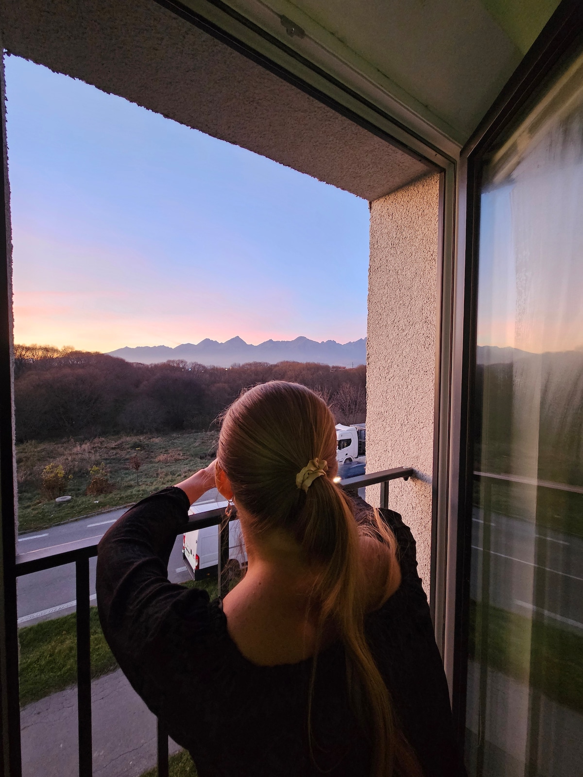 A view from a balcony frames a distant mountain range at sunset, with soft hues reflecting in the sky. A person is seen from behind, enjoying the scenery while leaning on the balustrade, creating a sense of relaxation in a natural setting.