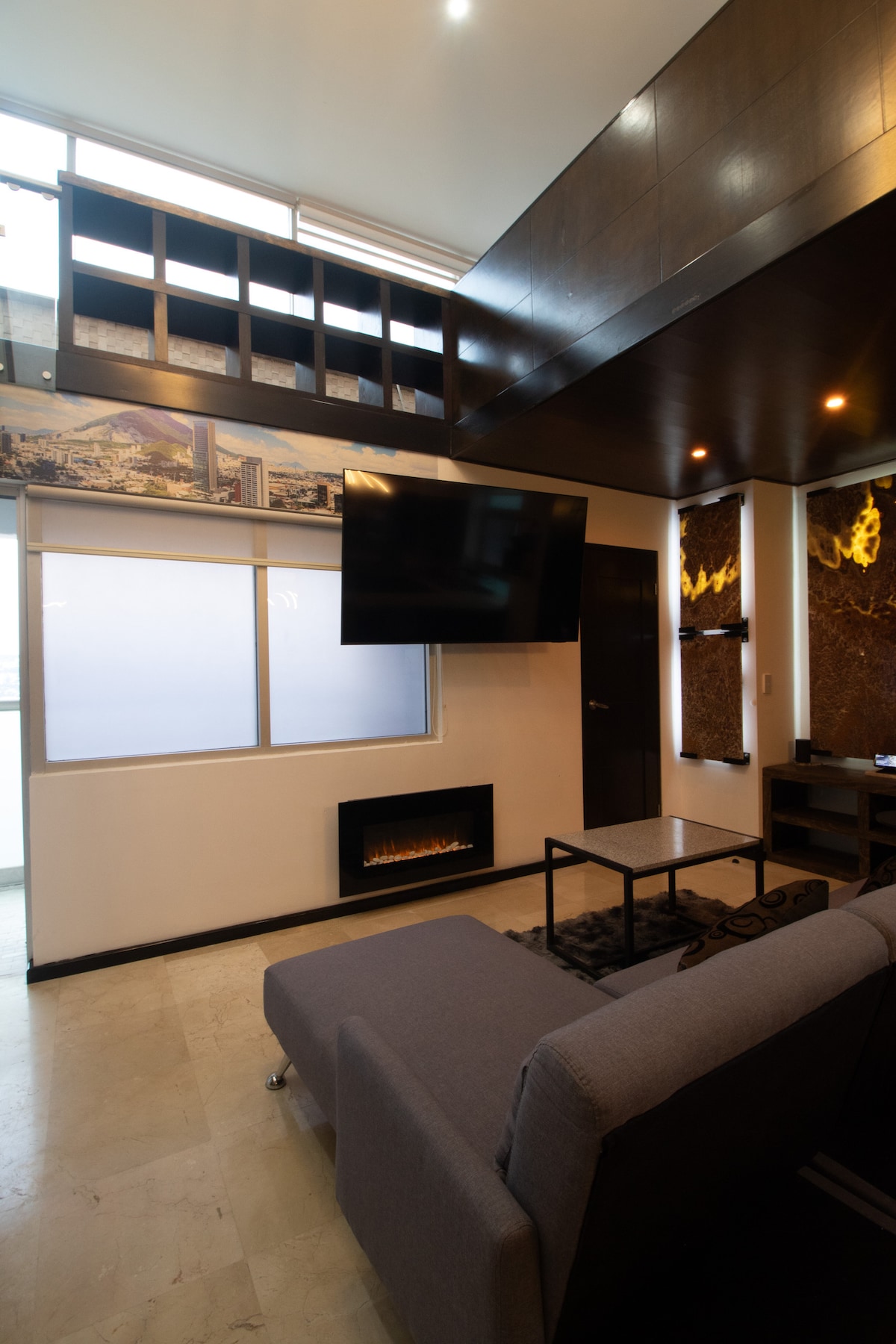 A modern living area is highlighted by a wall-mounted television and a contemporary electric fireplace. Comfortable seating is provided by a sofa, with a coffee table placed in front. Natural light enters through a large window, enhancing the room's spacious feel.