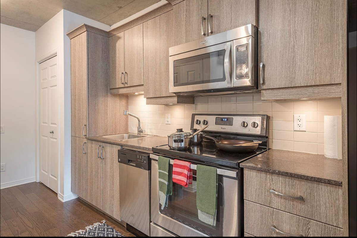 A modern kitchen is equipped with stainless steel appliances, including a microwave and oven. Light wood cabinetry complements the grey countertops. A small pot sits on the stovetop, while kitchen towels are draped over the oven handle. Soft lighting enhances the workspace.