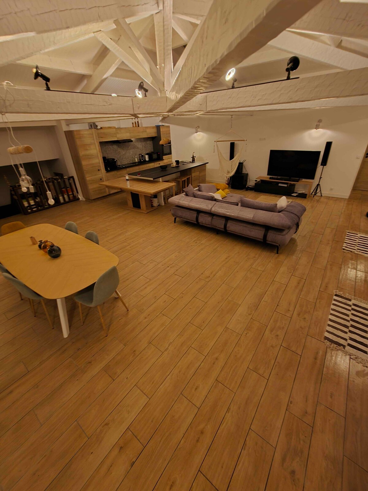 A spacious living area features a large sofa and a dining table with colorful chairs. The open kitchen is visible in the background, equipped with modern appliances. High ceilings and wooden flooring contribute to a welcoming environment, while a television is positioned near the seating area.