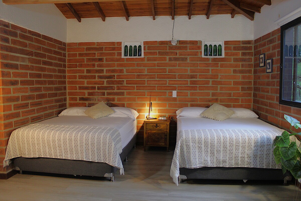 The inviting room features two spacious beds, each adorned with neutral bedding. A warm light emanates from a bedside table, while textured brick walls provide charm. A window allows natural light, and a small decorative plant adds a touch of greenery to the space.