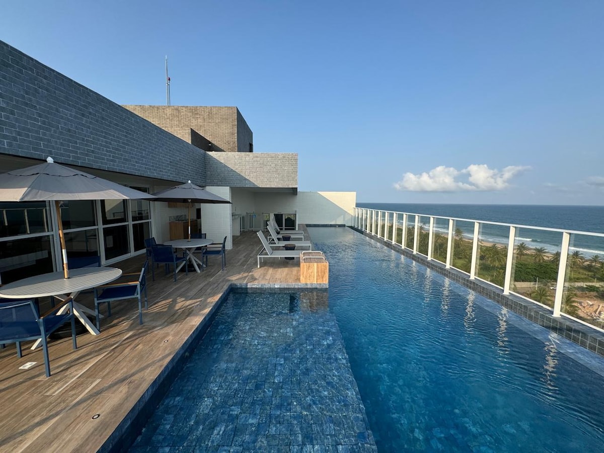 An outdoor pool area is depicted, featuring a shimmering blue pool with lounge chairs and umbrellas positioned nearby. The bright sky complements the tranquil ocean view, while a wooden deck enhances the space's inviting atmosphere.