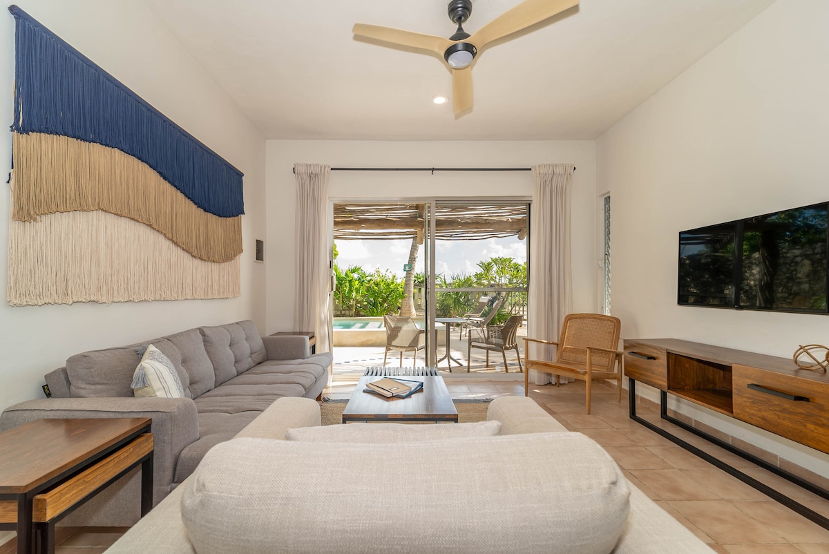 A light-filled living area features a comfortable sectional sofa and an armchair. Large glass doors lead to an outdoor space, allowing for natural light to brighten the room. A wall-mounted television and a wooden side table complete the inviting layout.